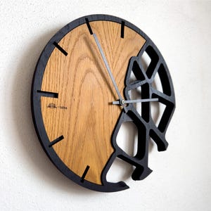 Può includere: Un orologio da parete circolare con un quadrante in legno color marrone chiaro naturale sul lato sinistro e un design geometrico a telaio aperto nero sul lato destro. Presenta sottili lancette in metallo nero e indici neri. Il testo tatra è stampato sul legno.
