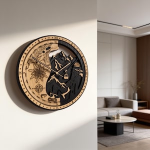 Op de afbeelding: Een ronde houten wandklok met een gespleten ontwerp, met aan de lichte houten kant een kompas, edelweissbloem en touwknoop, en aan de donkere houten kant een berglandschap, een kleine hut en een kabelbaan met wijzers.