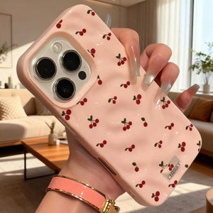 May include: A pink phone case adorned with a repeating pattern of red cherries and green stems. The case features a raised edge around the camera lenses and the word "CHERRY" printed in white. The case is held by a person.