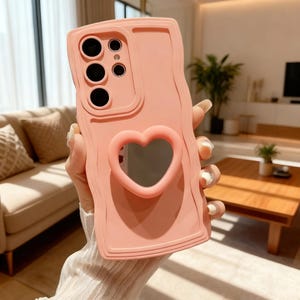 May include: A pale pink phone case featuring a heart-shaped aperture and a textured, wavy pattern. The case has cutouts for the phone's camera and buttons. The case is made of a soft material and is designed to protect the phone.
