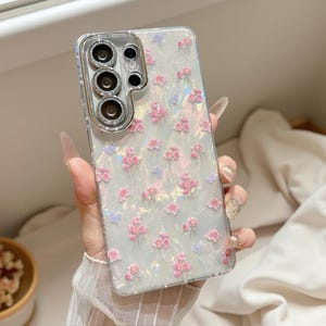 Puede incluir: Una funda transparente para smartphone con acabado iridiscente y nacarado, con un patrón repetitivo de pequeñas flores rosas y mariposas de color violeta claro. El área de la cámara tiene un borde plateado metálico con cristales incrustados.