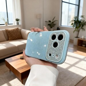 May include: A pale blue phone case with a glitter effect. The camera section is framed with small, sparkling rhinestones. The case is held in a person's hand.
