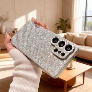 May include: A silver phone case adorned with shimmering rhinestones. The case features a camera lens protector with a silver surround. The phone case is held in a person's hand.