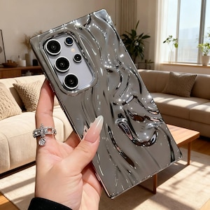 May include: A rectangular smartphone case with a highly reflective, metallic silver finish and a three-dimensional, liquid-like wavy texture. The case features a precise cutout for a multi-lens camera array, showcasing a sleek, modern aesthetic.