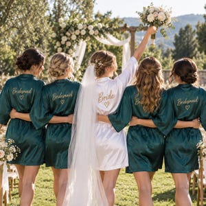 Könnte beinhalten: Fünf Personen stehen in einer Reihe und tragen Satin-Morgenmäntel. Vier Mäntel sind dunkel smaragdgrün mit dem Wort Bridesmaid und einem Herz in Gold gestickt. Der mittlere Mantel ist weiß mit dem Wort Bride und einem Herz in Gold.