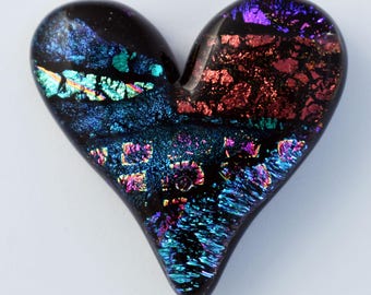 May include: A heart-shaped glass cabochon featuring a deep black base with iridescent dichroic inclusions. The surface displays vibrant, reflective patches of blue, teal, purple, and copper-red, creating a shimmering, multi-colored effect.