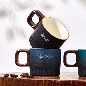 May include: Four ceramic mugs with names printed on them are displayed on a wooden surface. The mugs are in shades of blue, teal, and brown, with a dark brown handle. The mug in the front has the name "Josiah" printed on it.