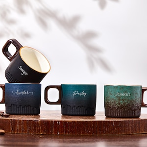 May include: Four ceramic mugs with dark brown square handles and textured bases. One black mug is tilted on top of a blue mug labeled "Hunter". A teal mug labeled "Dailey" and a green speckled mug labeled "Josiah" sit beside it. The black mug is labeled "Sawyer".