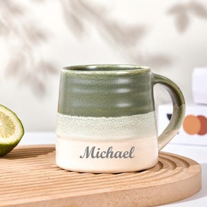 Op de afbeelding: Een keramische mok met een groen, wit en beige kleurenschema, gepersonaliseerd met de naam "Michael". De mok staat op een houten dienblad naast een halve limoen en een croissant. De mok heeft een handvat en een brede basis.