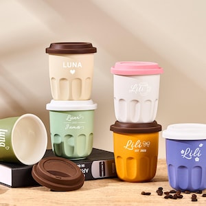 May include: A collection of colorful reusable coffee cups with lids. The cups are in various colors, including yellow, green, white, and purple, each with a name printed on them. Some cups are stacked, and one is lying on its side.