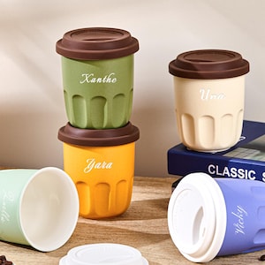 May include: A collection of five small, fluted cups in green, yellow, light green, cream, and purple, each featuring a personalized name in white script. The cups have brown or white lids, and some are stacked or lying on their sides.