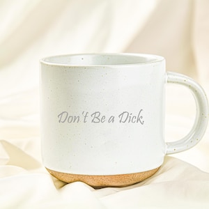 May include: A white ceramic mug with a speckled finish and a handle on the right side. The text "Don't Be a Dick." is printed in a dark gray script font on the front of the mug. The base of the mug is made of unglazed, light brown clay.