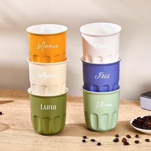 Könnte beinhalten: Sechs stapelbare Keramikbecher in Gelb, Creme, Olivgrün, Weiß, Blau und Hellgrün. Jeder Becher hat eine vertikal gerippte Textur am Boden und einen individuell aufgedruckten weißen Namen auf der Vorderseite: Mamie, Sophie, Luna, Chloe, Iris und Elona.