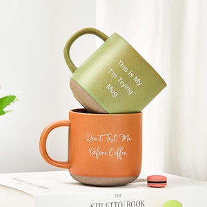 May include: Two ceramic mugs stacked vertically. The top mug is sage green with the text "This Is My 'I'm Trying' Mug". The bottom mug is terracotta orange with the text "Don't Test Me Before Coffee". Both mugs feature a speckled finish and a dark base.