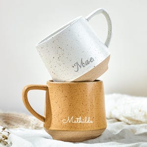 May include: Two stacked ceramic mugs with speckled finishes. The top mug is white with the name Mae printed in script. The bottom mug is mustard yellow with the name Mathilde printed in script. Both mugs feature a smooth handle and a natural base.