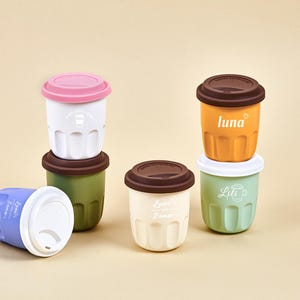 Puede incluir: Una colección de tazas de café reutilizables en varios colores, incluyendo blanco, naranja, verde y azul. Cada taza tiene una tapa a juego y una tapa marrón o rosa en contraste. Algunas tazas tienen texto, como "luna" y "Lili".