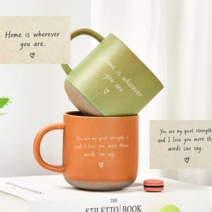 Può includere: Una tazza verde salvia impilata su una tazza arancione, entrambe con texture maculate e testo bianco. La tazza verde riporta la scritta "Home is wherever you are." La tazza arancione riporta "You are my quiet strength — and I love you more than words can say."