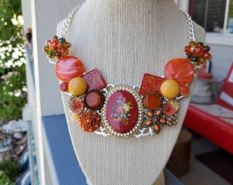 May include: A statement necklace with a mix of orange, red, and yellow buttons, beads, and a floral cameo. The necklace is on a white mannequin bust.