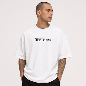 Pode incluir: T-shirt branco oversized com a frase "CHRIST IS KING" impressa em preto. A camisa tem mangas curtas e gola redonda. O modelo está usando calças pretas.