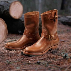 May include: A pair of mid-calf engineer boots crafted from smooth tan leather. These boots feature a rounded toe, a stacked heel, and two adjustable straps with gold-toned metal buckles located at the ankle and the upper shaft opening.