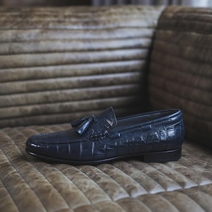 Peut inclure: Un mocassin en cuir bleu marine foncé avec un motif texturé en peau de crocodile, un bout arrondi et un détail à pompons sur l'empeigne. La chaussure présente un petit talon et une semelle sombre lisse, offrant une finition polie et structurée.