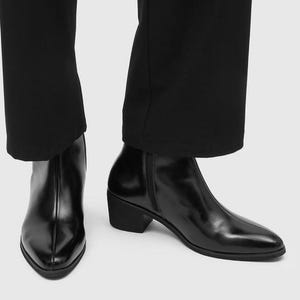 Könnte beinhalten: Ein Paar schwarze Stiefeletten mit polierter Oberfläche, spitzer Zehenpartie, einer durchgehenden Mittelnaht auf der Vorderseite und einem Blockabsatz. Die Stiefel haben einen seitlichen Reißverschluss und werden zu einer schwarzen Hose getragen.