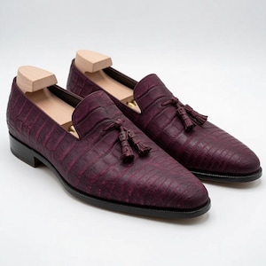 Può includere: Un paio di mocassini color bordeaux scuro con una finitura in pelle testurizzata effetto coccodrillo, una nappa decorativa sulla tomaia e una suola nera. Ogni scarpa include un tendiscarpe in legno chiaro per mantenerne la forma.