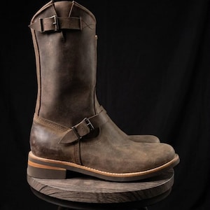 May include: A mid-calf brown leather boot featuring a rounded toe, a stacked heel, and two adjustable straps with metal buckles. One strap is positioned across the ankle area, while the second strap is located near the top of the boot shaft.