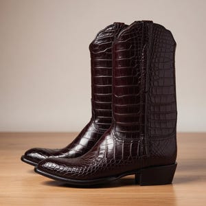Puede incluir: Un par de botas vaqueras de color marrón oscuro con una textura brillante en relieve de piel de cocodrilo. Las botas tienen punta puntiaguda, caña a media pantorrilla con borde superior festoneado y un tacón de bloque apilado. El acabado es brillante.