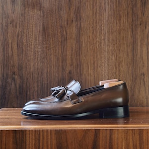 Peut inclure: Un mocassin en cuir marron foncé doté d'un détail à pampilles décoratif sur l'empeigne, d'un bout cousu et d'un petit talon noir empilé. Un embauchoir en bois clair est inséré à l'arrière de la chaussure pour conserver sa forme.