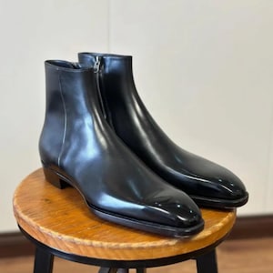 Puede incluir: Un par de botines de cuero negro con un acabado liso y pulido, punta puntiaguda, tacón bajo de bloque y cierre de cremallera lateral vertical. Los botines presentan una silueta limpia y aerodinámica con costuras mínimas y una suela negra.