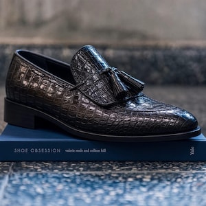 Può includere: Un mocassino marrone scuro con una texture lucida goffrata a coccodrillo, caratterizzato da una nappa decorativa sulla tomaia. La scarpa poggia su un libro blu intitolato SHOE OBSESSION di Valerie Steele e Colleen Hill, pubblicato da Yale.