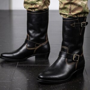 May include: A pair of black leather mid-calf boots featuring a rounded toe, a stacked heel, and contrasting tan stitching. Each boot includes a decorative strap across the instep and an adjustable buckle strap at the top, both secured with metal hardware.
