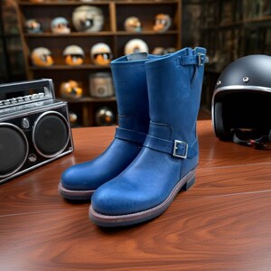 May include: A pair of mid-calf blue leather boots featuring a rounded toe, a stacked heel, and a silver-toned buckle strap across the vamp. The boots have a secondary adjustable strap with a silver buckle at the top of the shaft and a dark sole.
