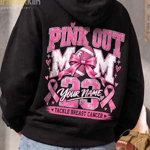 Puede incluir: Una sudadera negra con capucha que presenta un gráfico en la parte posterior con el texto PINK OUT MOM 23, un balón de fútbol americano con un lazo rosa y cintas de concienciación rosas. El diseño incluye el texto YOUR NAME y TACKLE BREAST CANCER.