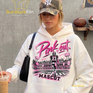 Puede incluir: Una sudadera con capucha blanca que presenta un estampado gr&aacute;fico de un jugador de f&uacute;tbol americano en un campo con el texto Pink out y MASCOT. El dise&ntilde;o incluye cintas rosas, luces de estadio y un jugador con el n&uacute;mero 23.