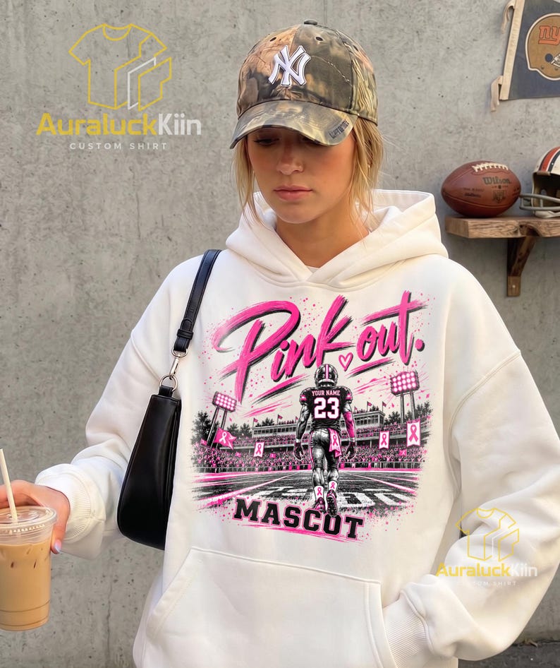 Puede incluir: Una sudadera con capucha blanca que presenta un estampado gr&aacute;fico de un jugador de f&uacute;tbol americano en un campo con el texto Pink out y MASCOT. El dise&ntilde;o incluye cintas rosas, luces de estadio y un jugador con el n&uacute;mero 23.