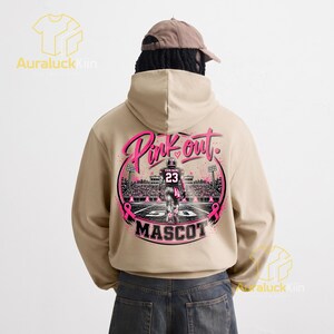 Puede incluir: Una sudadera con capucha de color beige que presenta un gr&aacute;fico en la parte posterior. El dise&ntilde;o muestra a un jugador de f&uacute;tbol americano en un campo con el texto Pink out y MASCOT. Incluye lazos rosas, luces de estadio y una camiseta con el n&uacute;mero 23.