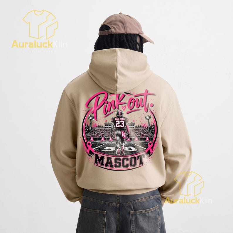 Puede incluir: Una sudadera con capucha de color beige que presenta un gr&aacute;fico en la parte posterior. El dise&ntilde;o muestra a un jugador de f&uacute;tbol americano en un campo con el texto Pink out y MASCOT. Incluye lazos rosas, luces de estadio y una camiseta con el n&uacute;mero 23.