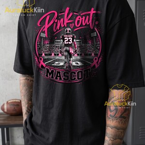 Puede incluir: Una camiseta negra con un gr&aacute;fico de un jugador de f&uacute;tbol americano en un campo con el texto Pink out y MASCOT. El dise&ntilde;o incluye cintas rosas, luces de estadio y un jugador con una camiseta con el n&uacute;mero 23 y el texto Your Name.