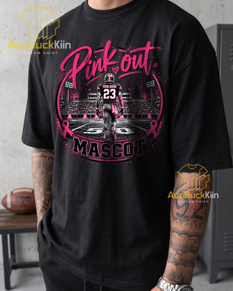 Puede incluir: Una camiseta negra con un gr&aacute;fico de un jugador de f&uacute;tbol americano en un campo con el texto Pink out y MASCOT. El dise&ntilde;o incluye cintas rosas, luces de estadio y un jugador con una camiseta con el n&uacute;mero 23 y el texto Your Name.