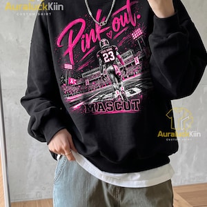 Puede incluir: Una sudadera negra de manga larga con un estampado gr&aacute;fico de un jugador de f&uacute;tbol americano en un campo con el texto Pink out y Mascot. El dise&ntilde;o incluye detalles en rosa, luces de estadio y s&iacute;mbolos de lazos. Tiene cuello redondo.