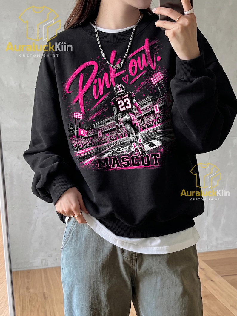 Puede incluir: Una sudadera negra de manga larga con un estampado gr&aacute;fico de un jugador de f&uacute;tbol americano en un campo con el texto Pink out y Mascot. El dise&ntilde;o incluye detalles en rosa, luces de estadio y s&iacute;mbolos de lazos. Tiene cuello redondo.