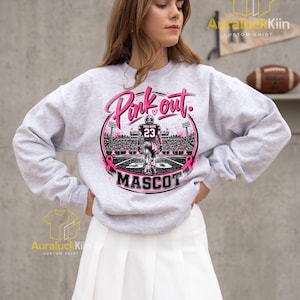 Puede incluir: Una sudadera gris claro de cuello redondo con un estampado gr&aacute;fico de un jugador de f&uacute;tbol americano en un campo con el texto Pink out y MASCOT. El dise&ntilde;o incluye cintas rosas, luces de estadio y un jugador con el n&uacute;mero 23 en la camiseta.