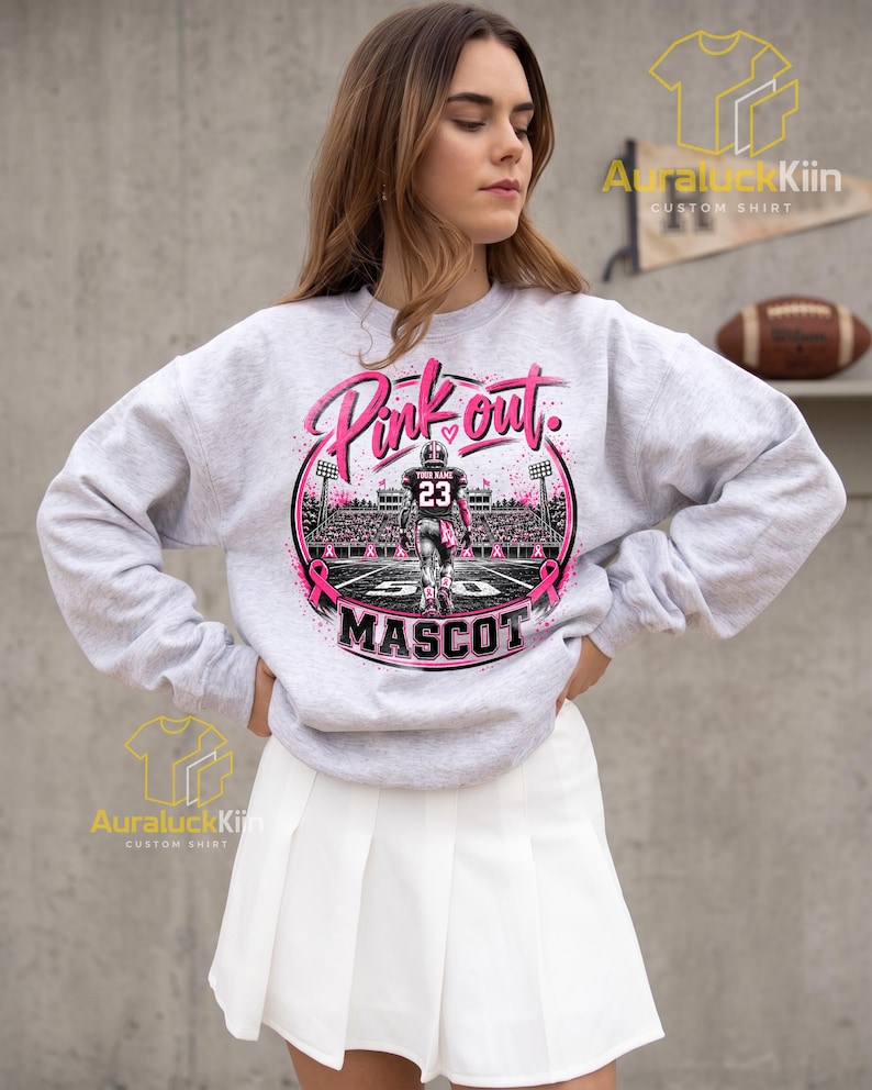Puede incluir: Una sudadera gris claro de cuello redondo con un estampado gr&aacute;fico de un jugador de f&uacute;tbol americano en un campo con el texto Pink out y MASCOT. El dise&ntilde;o incluye cintas rosas, luces de estadio y un jugador con el n&uacute;mero 23 en la camiseta.