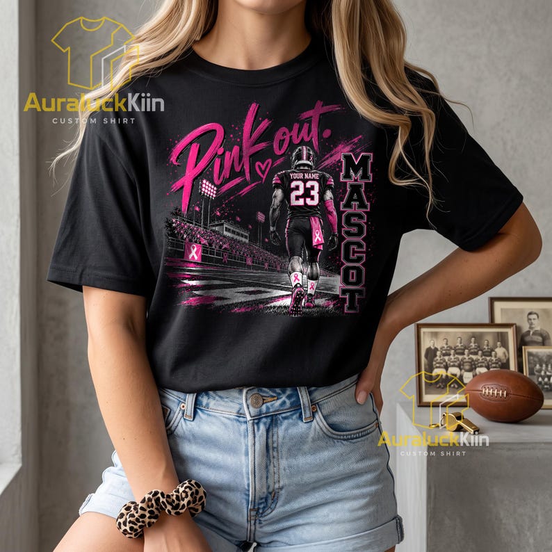 Puede incluir: Una camiseta negra de manga corta con un gr&aacute;fico de un jugador de f&uacute;tbol americano con el n&uacute;mero 23 y el texto "Pink out" y "MASCOT". El dise&ntilde;o incluye detalles en rosa, un fondo de estadio y un s&iacute;mbolo de coraz&oacute;n.