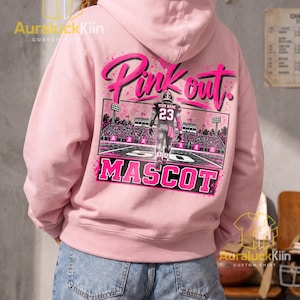 Puede incluir: Una sudadera con capucha de color rosa claro que presenta un gr&aacute;fico en la parte posterior con el texto Pink out y MASCOT. El dise&ntilde;o incluye un jugador de f&uacute;tbol americano con el n&uacute;mero 23 en un campo con luces y lazos rosas en las gradas.