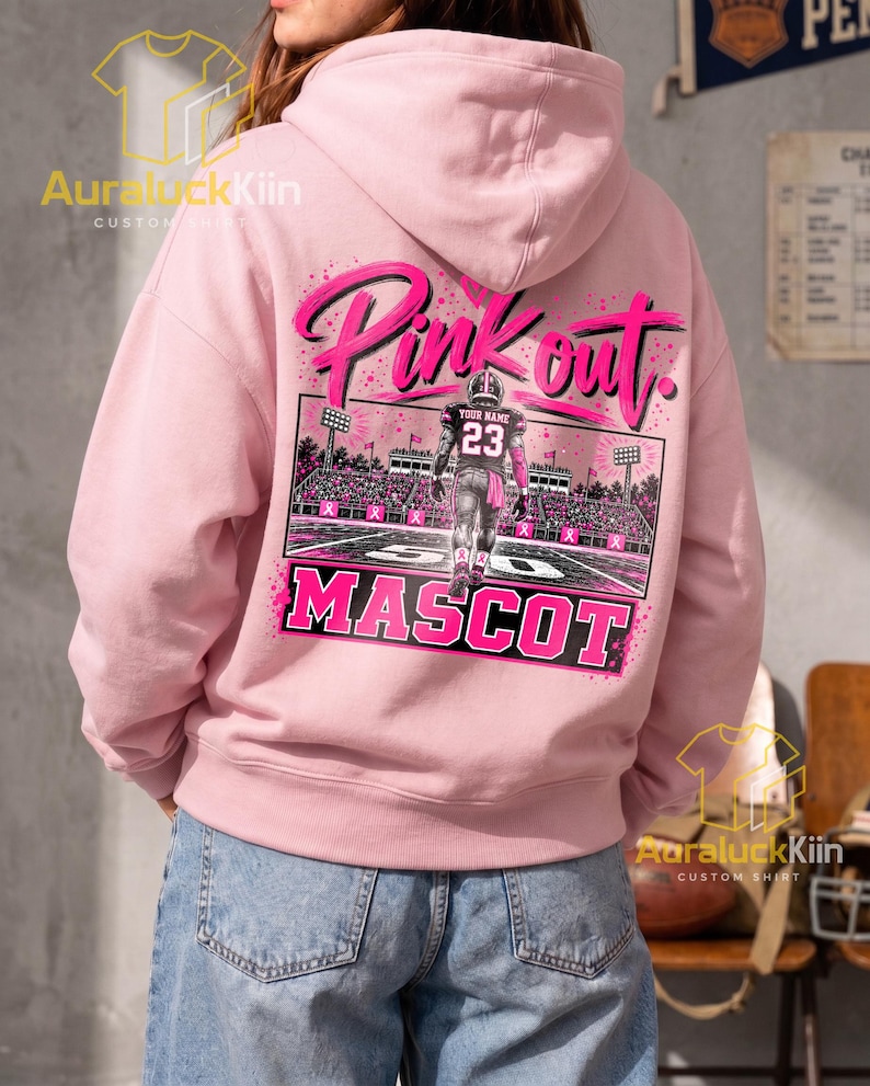 Puede incluir: Una sudadera con capucha de color rosa claro que presenta un gr&aacute;fico en la parte posterior con el texto Pink out y MASCOT. El dise&ntilde;o incluye un jugador de f&uacute;tbol americano con el n&uacute;mero 23 en un campo con luces y lazos rosas en las gradas.