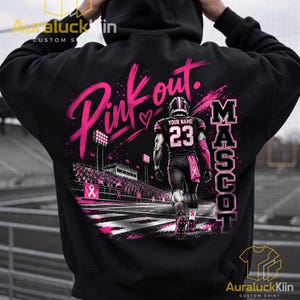 Puede incluir: Una sudadera negra con capucha que presenta un gr&aacute;fico en la espalda de un jugador de f&uacute;tbol americano con el n&uacute;mero 23 y el texto YOUR NAME. El dise&ntilde;o incluye el texto Pink out y MASCOT, con detalles en rosa e im&aacute;genes de un estadio.