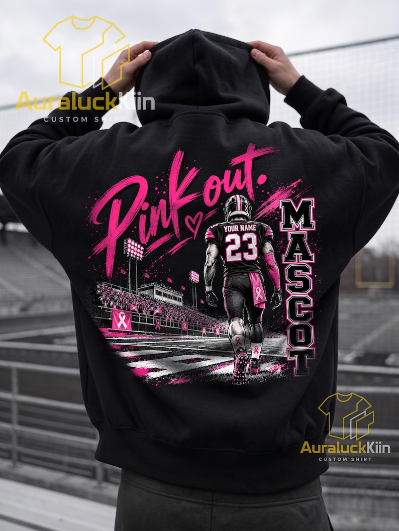 Puede incluir: Una sudadera negra con capucha que presenta un gr&aacute;fico en la espalda de un jugador de f&uacute;tbol americano con el n&uacute;mero 23 y el texto YOUR NAME. El dise&ntilde;o incluye el texto Pink out y MASCOT, con detalles en rosa e im&aacute;genes de un estadio.